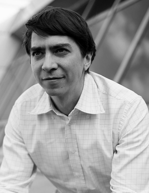 Дмитрий Сотников, чёрно-белое фото: сидит на крыше у остеклённого фасада, рубашка в клетку, руки сцеплены